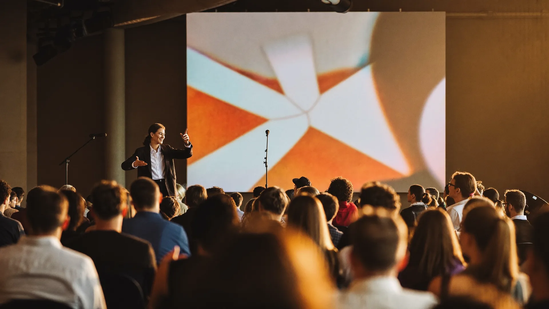 Speaker on a keynote stage before an audience, with a large screen in warm tones